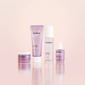 The Solawave Solabiome Glowing Skin Starter Kit’s four lavender-hued products are lined up on a glossy pale pink surface with a soft pink gradient backdrop.